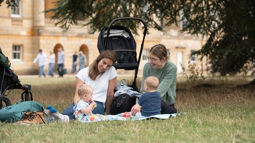 Two adults sit on a blanket on a lawn with two young children, playing with toys. A stroller, bag, and shoes are nearby, and a large stone building with arched windows is visible in the background beneath tree branches.